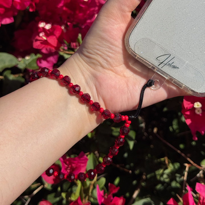Hand holding a phone with a cherry red beaded phone charm in front of pink flowers
