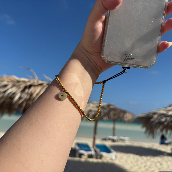 Hand holding a phone. The Golden Hatossa phone Charm is wrapped around the wrist. Background includes a beach.